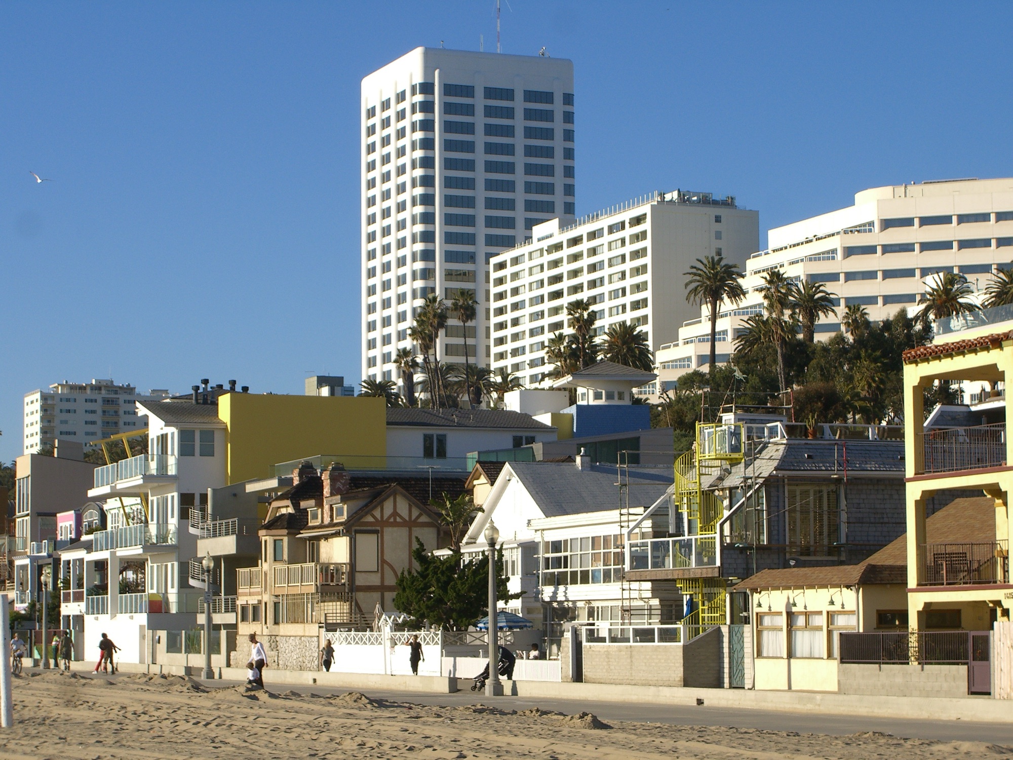 Santa Monica beachfront: a row of small two- and three-storey beach houses in front of a white high-rise office tower and large hotel blocks on the bluff above.