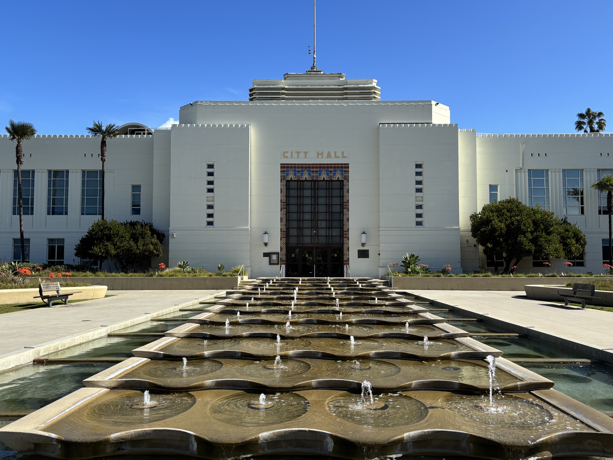 The white Art Deco facade of Santa Monica City Hall behind a long tiled reflecting fountain, with palm trees on either side.