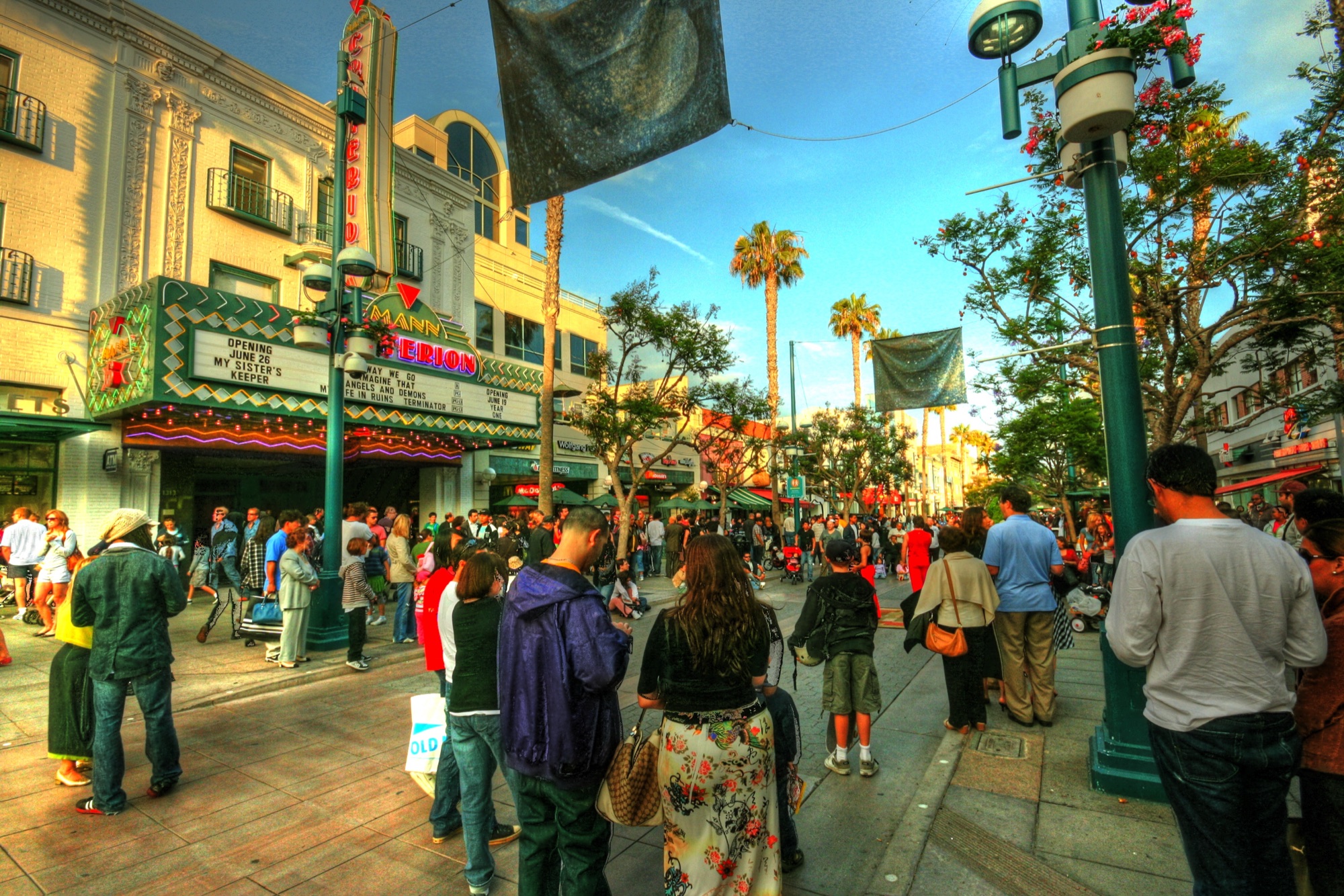 A crowded pedestrian street in the evening, with people filling the Promenade outside an Art Deco cinema whose neon marquee lists current films.