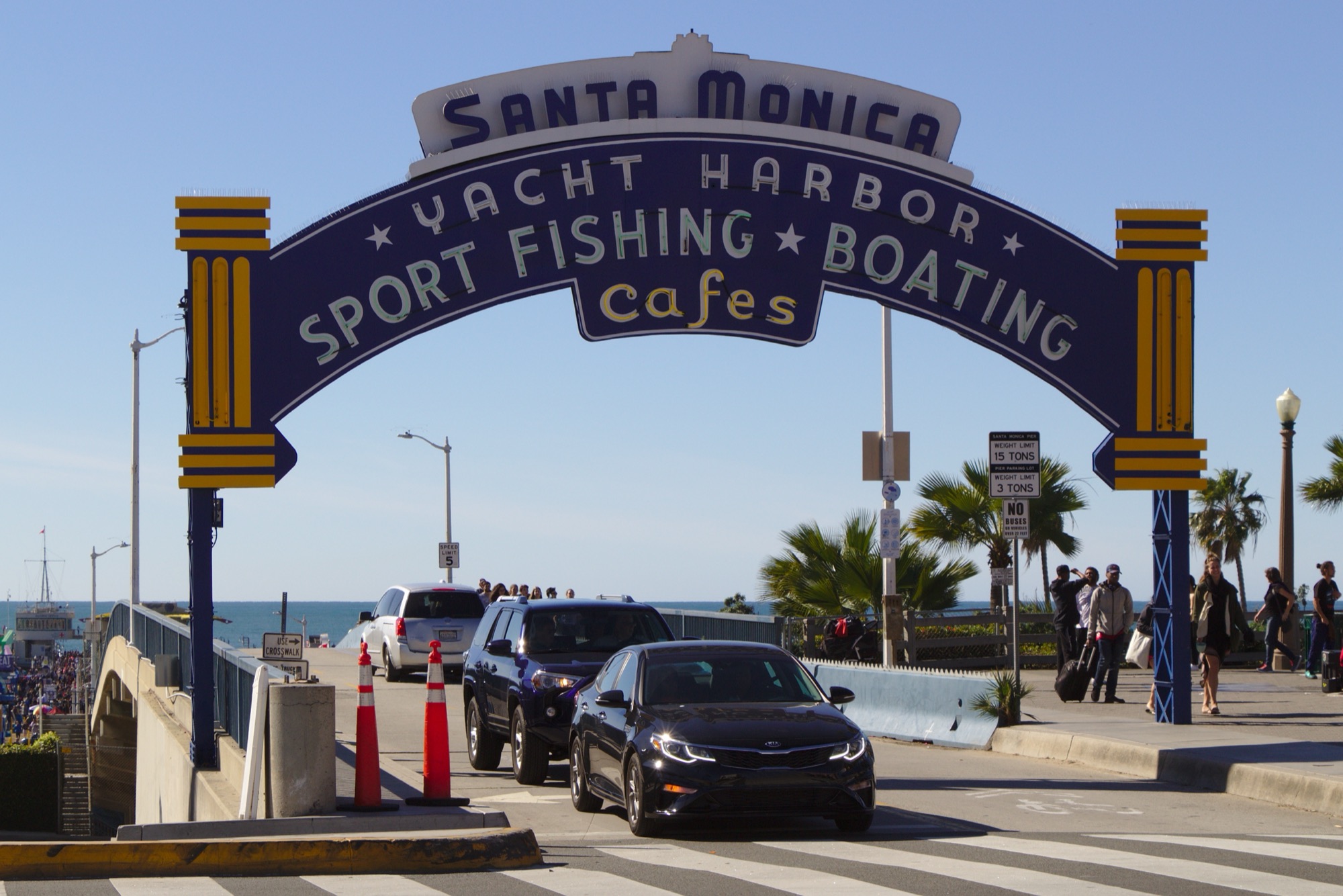 A large blue and yellow neon sign arching over a roadway at the entrance to the Santa Monica Pier, with cars passing beneath it and the ocean visible beyond.
